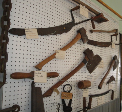 Carpentry Hand Tools.
Clockwise from top right: log scaler, hay knife, broad axe (used to square timber), hatchel, draw knife (to work wood), hand sander for floors, hay knife, axe, broad axe, wire tightener, wooden hammer, hay knife, scale, bridle chain (brakes on a sleigh when going down a steep hill). The chisel-edged broad axe was a particularly essential tool for the early settlers. A kind of plane or striking chisel used for hewing round logs into square beams, it was twice the size of a felling axe 